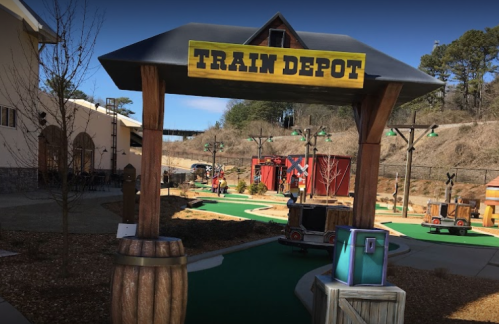 A miniature golf course entrance with a sign reading "Train Depot" and themed decorations in a scenic outdoor setting.