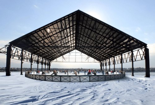 A large, open pavilion on a snowy landscape, with people skating on an ice rink beneath a bright sky.