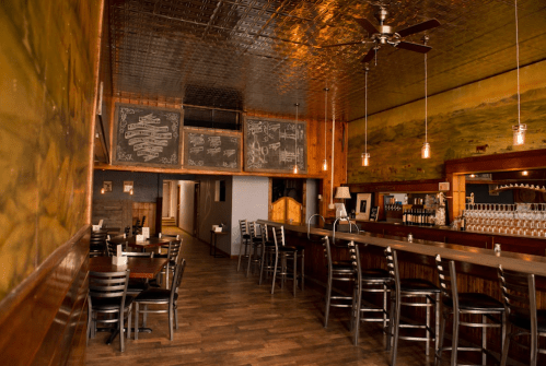 Interior of a rustic restaurant featuring wooden decor, a bar, and chalkboard menus under a tin ceiling.