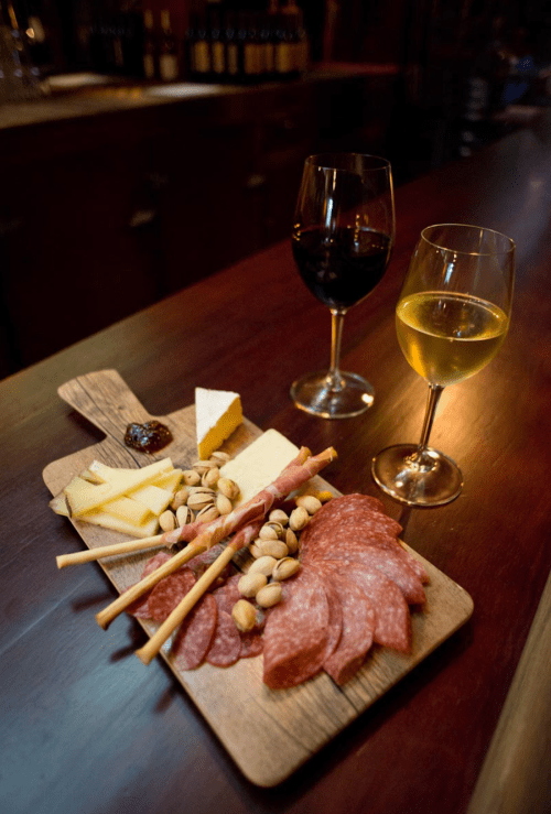 A wooden platter with assorted cheeses, salami, nuts, and breadsticks, accompanied by red and white wine glasses.