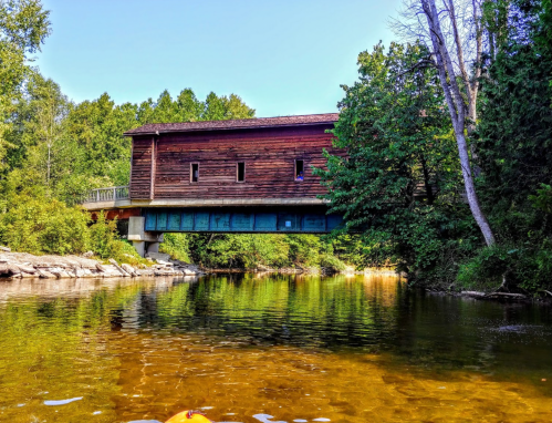 A wooden covered bridge spans a calm river, surrounded by lush greenery and trees under a clear blue sky.