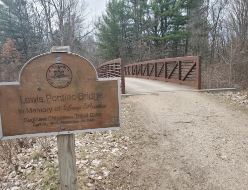 Sign for Lewis Pontiac Bridge, honoring Lewis Pontine, with a wooden bridge and trees in the background.