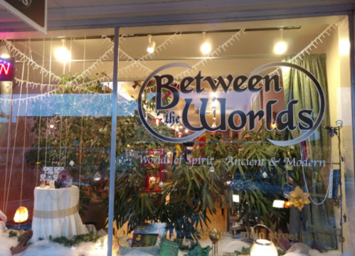Storefront of "Between the Worlds," featuring plants, decorative lights, and a sign promoting spiritual and modern items.