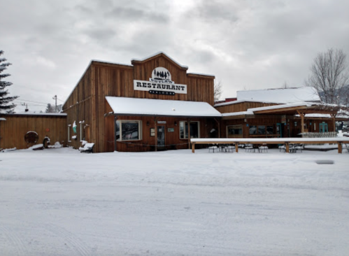 A rustic wooden restaurant building covered in snow, with outdoor seating and a cloudy sky above.
