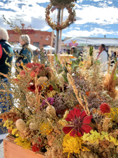 A vibrant display of dried flowers and foliage in a market setting, with people and a blue sky in the background.