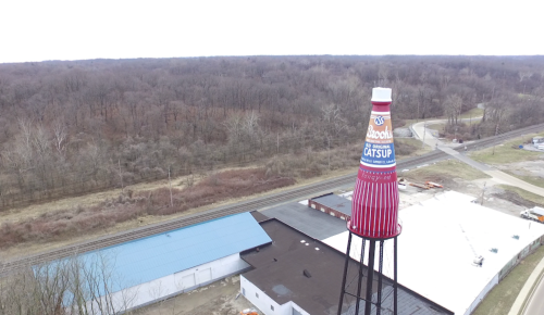 A large ketchup bottle water tower stands over a building near a wooded area and a road.