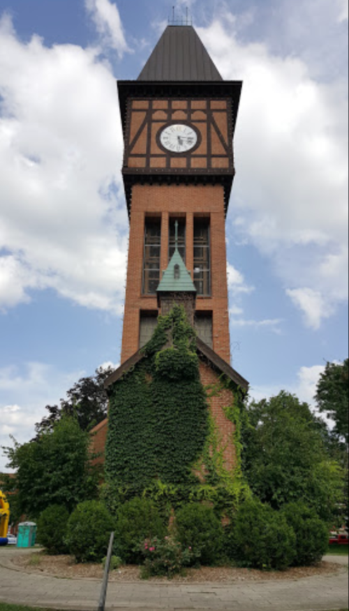 A tall clock tower with a triangular roof, surrounded by greenery and blue skies with scattered clouds.