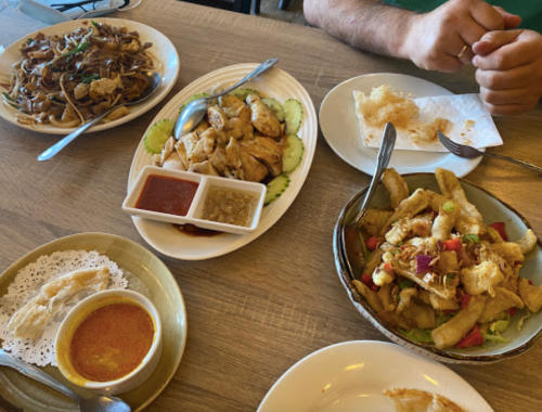A table with various dishes: noodles, chicken with sauces, a salad, and a dessert, with hands reaching for the food.