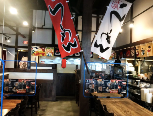 Interior of a Japanese restaurant featuring banners, wooden beams, and a visible kitchen area.