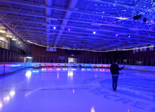 A person walks on an ice rink illuminated by colorful lights, with seating along the edge.