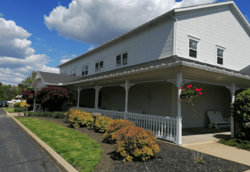 A white building with a porch, surrounded by colorful shrubs and flowers, under a partly cloudy sky.