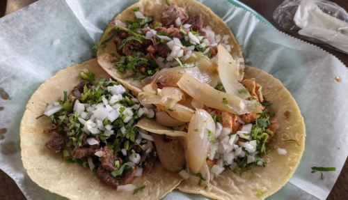 Three tacos filled with meat, topped with onions and chopped cilantro, served on a light blue paper.