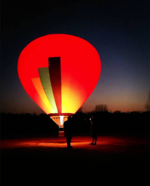 A brightly lit hot air balloon in red and green against a twilight sky, with a person standing nearby.
