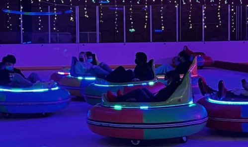 A group of people in colorful bumper cars on an ice rink, illuminated by vibrant lights and festive decorations.
