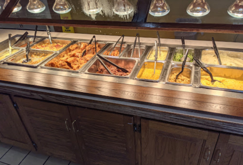 A buffet with various food trays, including fried chicken, macaroni and cheese, and vegetables, under warming lights.