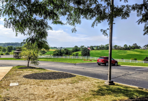 A red car parked on a paved area with green fields and trees in the background under a clear sky.