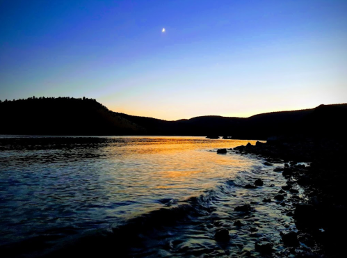 A serene twilight scene over a calm lake, with silhouettes of hills and a crescent moon in the sky.