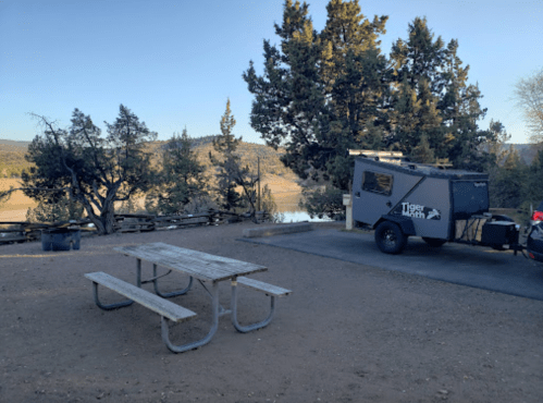 A campsite with a picnic table, a trailer, and trees overlooking a lake and mountains in the background.