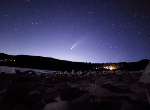 A comet streaks across a starry night sky above a dark landscape and a calm body of water.