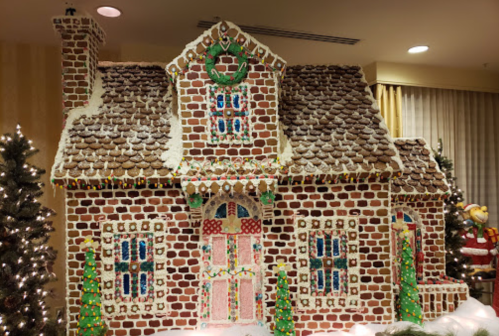 A large gingerbread house decorated with icing, candy, and festive greenery, surrounded by Christmas trees.