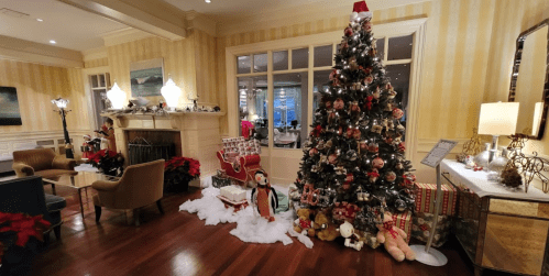 A cozy room decorated for Christmas, featuring a large tree, gifts, and festive ornaments by a fireplace.