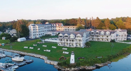 Aerial view of a waterfront hotel with white buildings, green lawns, and boats docked nearby, surrounded by trees.