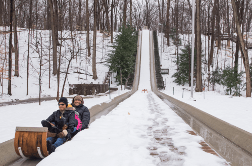 A family enjoys a snowy day on a sledding hill, with trees in the background and a clear path down the slope.