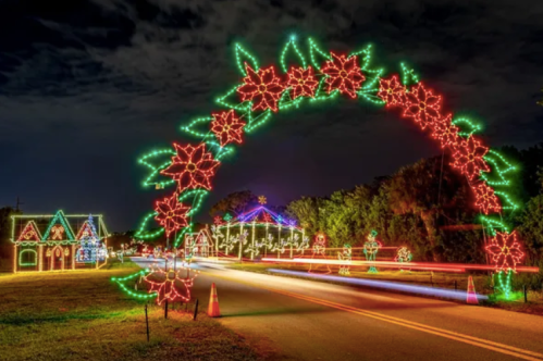 Colorful holiday lights form an arch over a road, with decorated houses and trees in the background against a night sky.