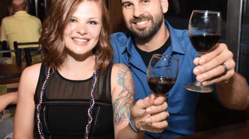 A smiling couple raises glasses of red wine, celebrating together in a lively setting.
