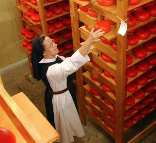 A nun reaches for a round cheese on a wooden shelf filled with red cheeses in a storage room.