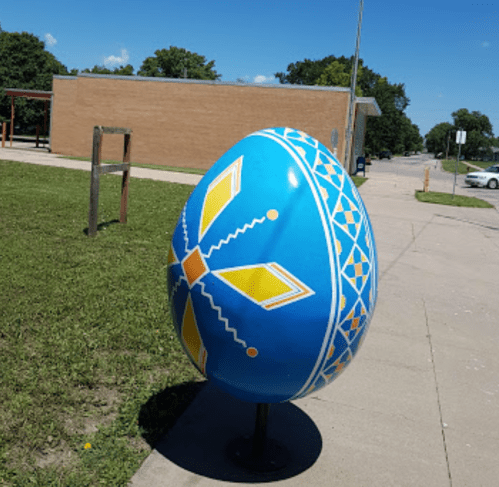 A large, colorful decorative egg with blue and yellow patterns stands on a sidewalk in a grassy area.