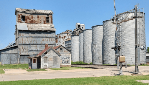 Abandoned industrial buildings and grain silos under a clear blue sky, with a small house in the foreground.