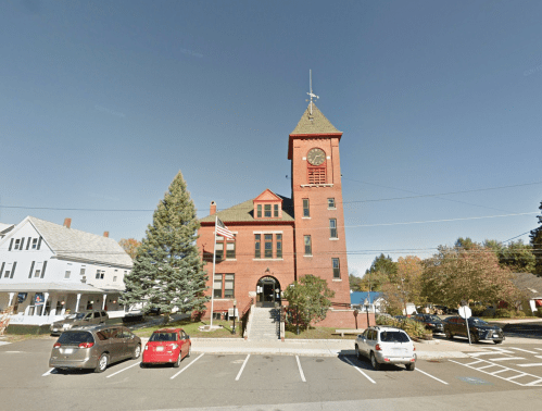 Historic brick building with a clock tower, surrounded by trees and parked cars, under a clear blue sky.
