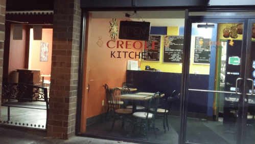 Exterior view of Creole Kitchen at night, featuring a "Open" sign and colorful walls with tables visible inside.