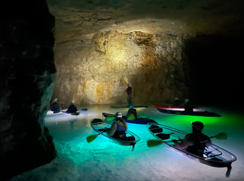 A group of kayakers paddles in a dimly lit cave, illuminated by colorful lights reflecting off the water.