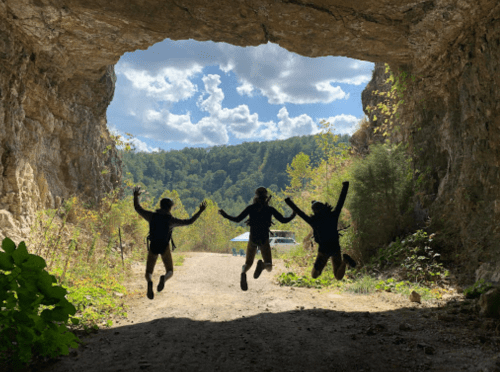Three silhouetted figures jump joyfully in front of a cave opening, with lush greenery and a blue sky in the background.