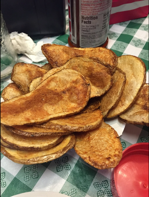 A pile of golden-brown potato chips on a checkered tablecloth, with a bottle of ketchup in the background.