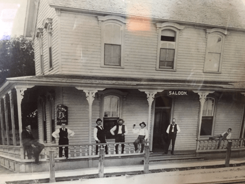 Historic photo of a saloon with six people posing on the porch of a wooden building.