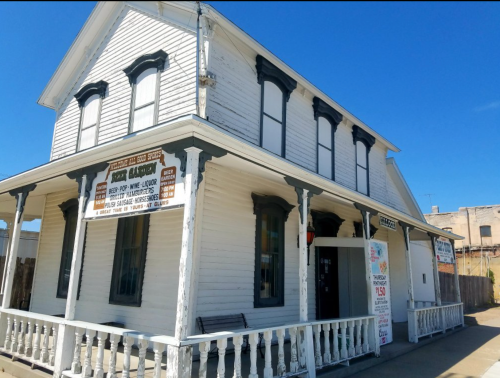 A historic white building with a porch, featuring signs for a general store and liquor, under a clear blue sky.
