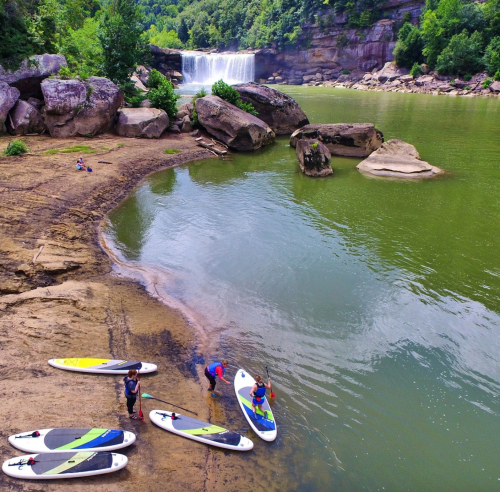 A serene river scene with paddleboarders near a waterfall, surrounded by lush greenery and rocky terrain.