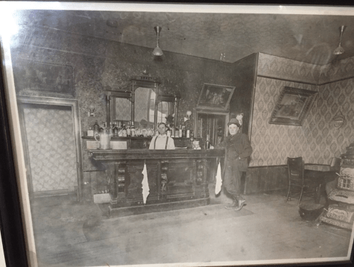 A vintage black-and-white photo of two men in a bar, with a wooden counter and ornate wallpaper.