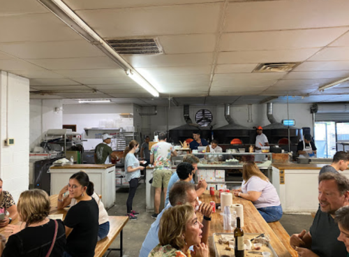 A busy restaurant scene with diners at wooden tables and staff preparing food in the kitchen.