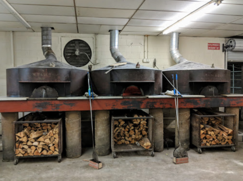Three large ovens with chimneys, above wooden racks stacked with firewood, in a well-lit industrial kitchen.