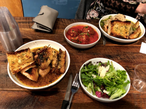 A wooden table with three bowls of food: grilled chicken, meatballs in sauce, and a fresh salad.