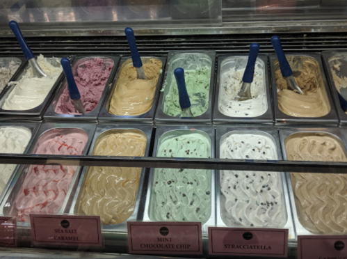A display of various gelato flavors in metal tubs, including mint chocolate chip and stracciatella, with blue scoops.
