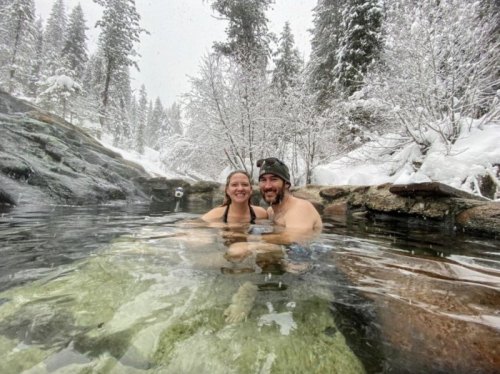 A couple smiles while relaxing in a natural hot spring surrounded by snowy trees and a winter landscape.