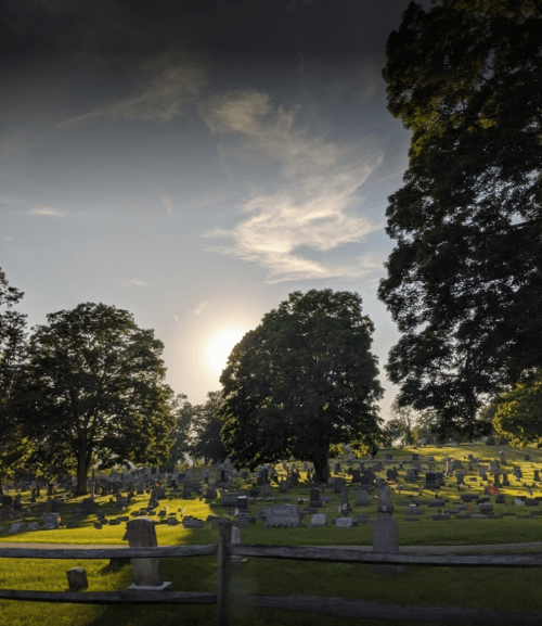 A serene cemetery at sunset, with gravestones scattered among lush green trees and grass.