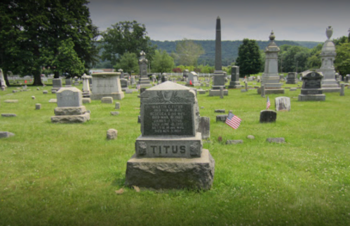 A cemetery scene featuring a gravestone marked "TITUS" among other headstones and flags on a grassy landscape.