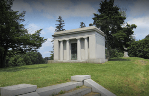 A stone mausoleum with columns, set on a grassy hill surrounded by trees under a partly cloudy sky.