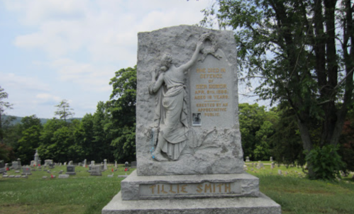 A gravestone with a carved figure and inscription for Tillie Smith, located in a cemetery with trees in the background.